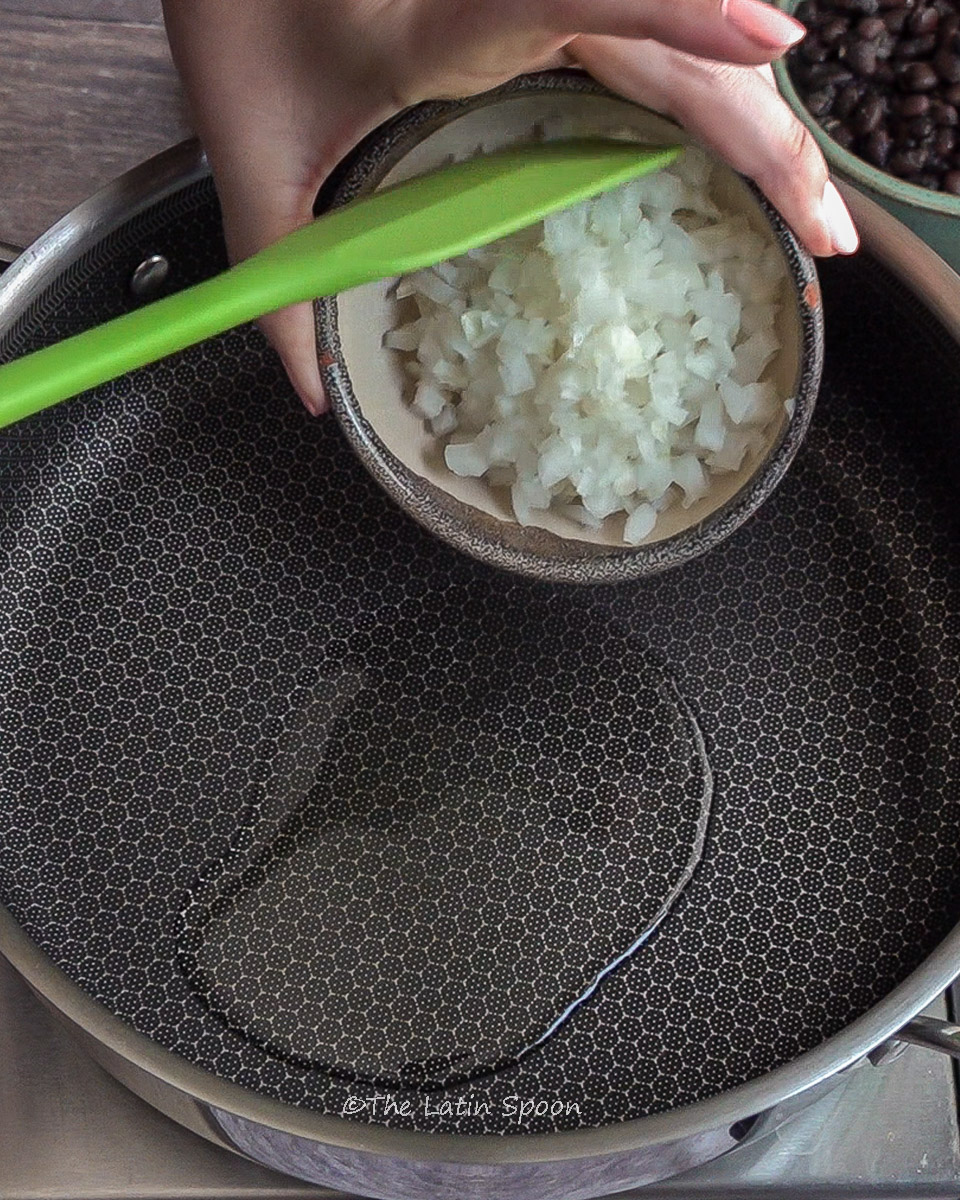 Large saucepan or skillet with a little oil. Above it, a left hand holds a small bowl of onions, being pushed out of the bowl with a green spatula.