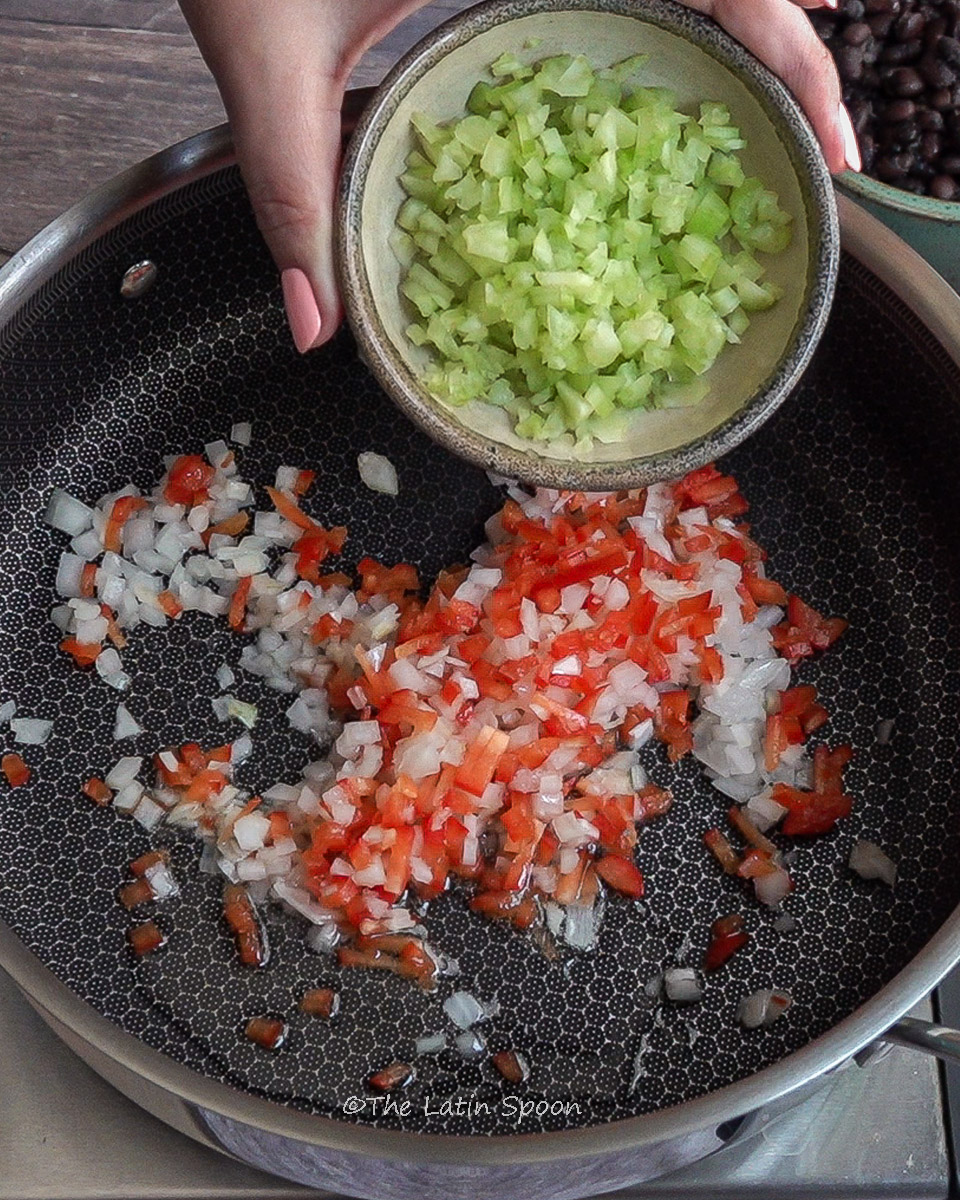 Large pan with chopped onions and bell peppers cooking in oil, while a left hand holds a small bowl of chopped celery, showing the next step of adding celery to the pan.