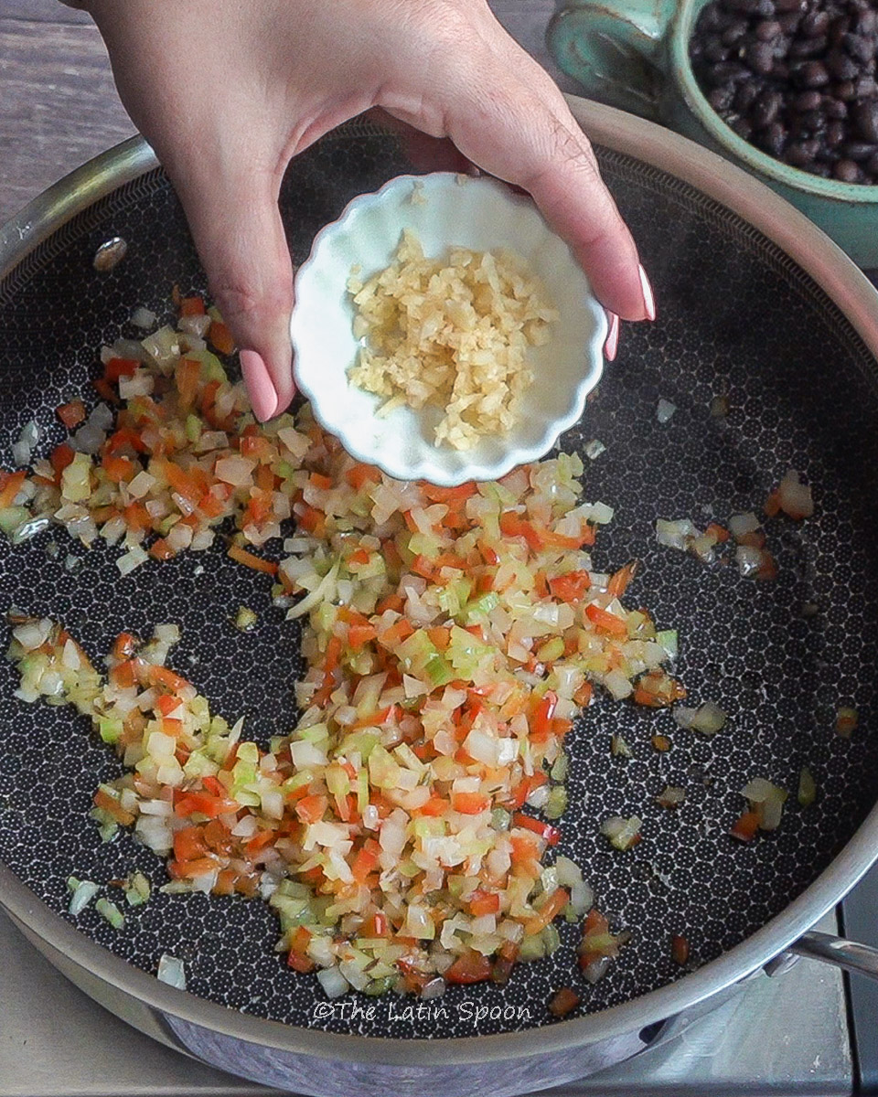 Pan with sofrito (onions, celery, bell peppers, and oil) cooking, while the left hand is about to add minced garlic from a small bowl.