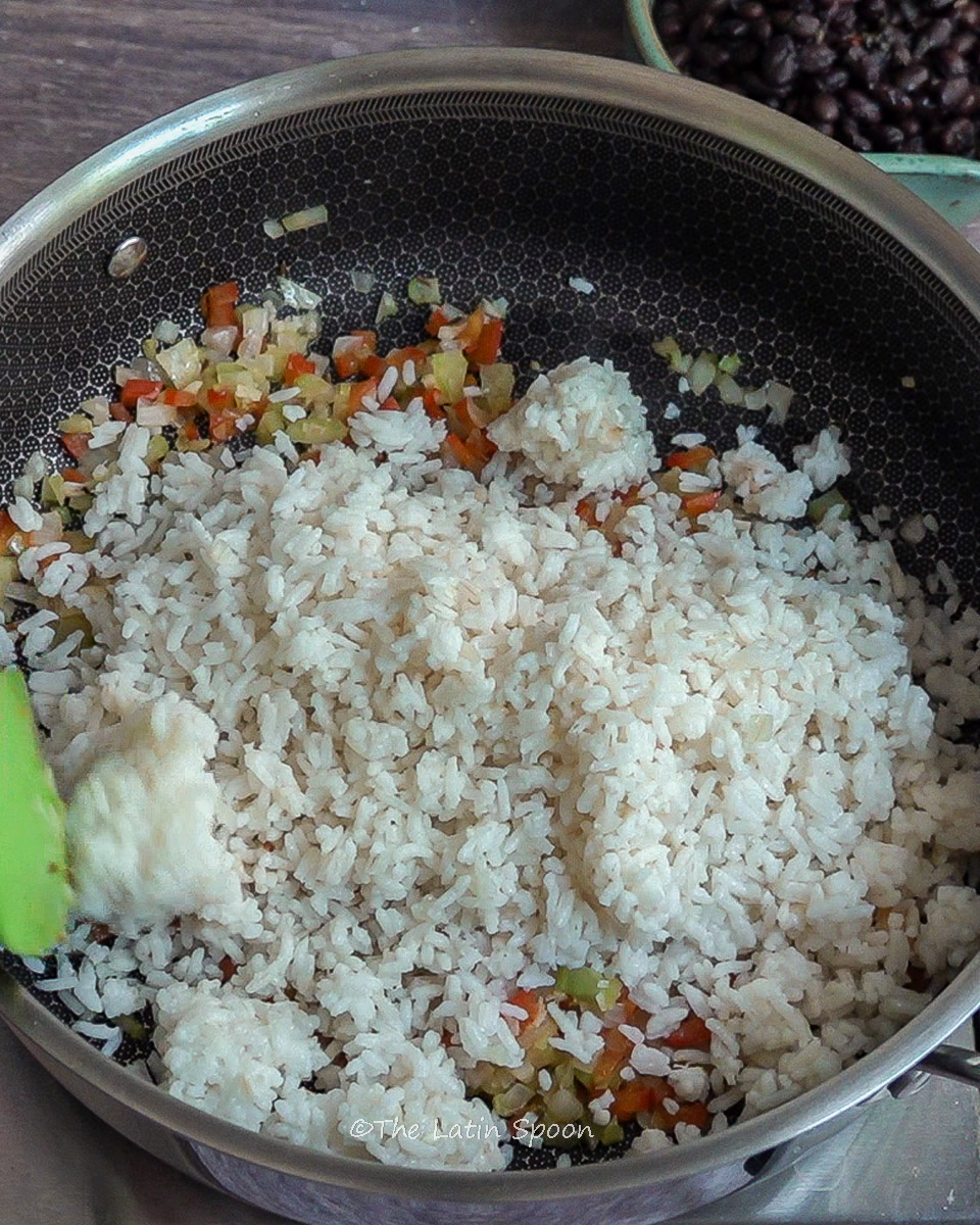 Large pan showing sofrito at the bottom with fully cooked white rice added on top. In the corner, a green spatula begins to mix the ingredients.