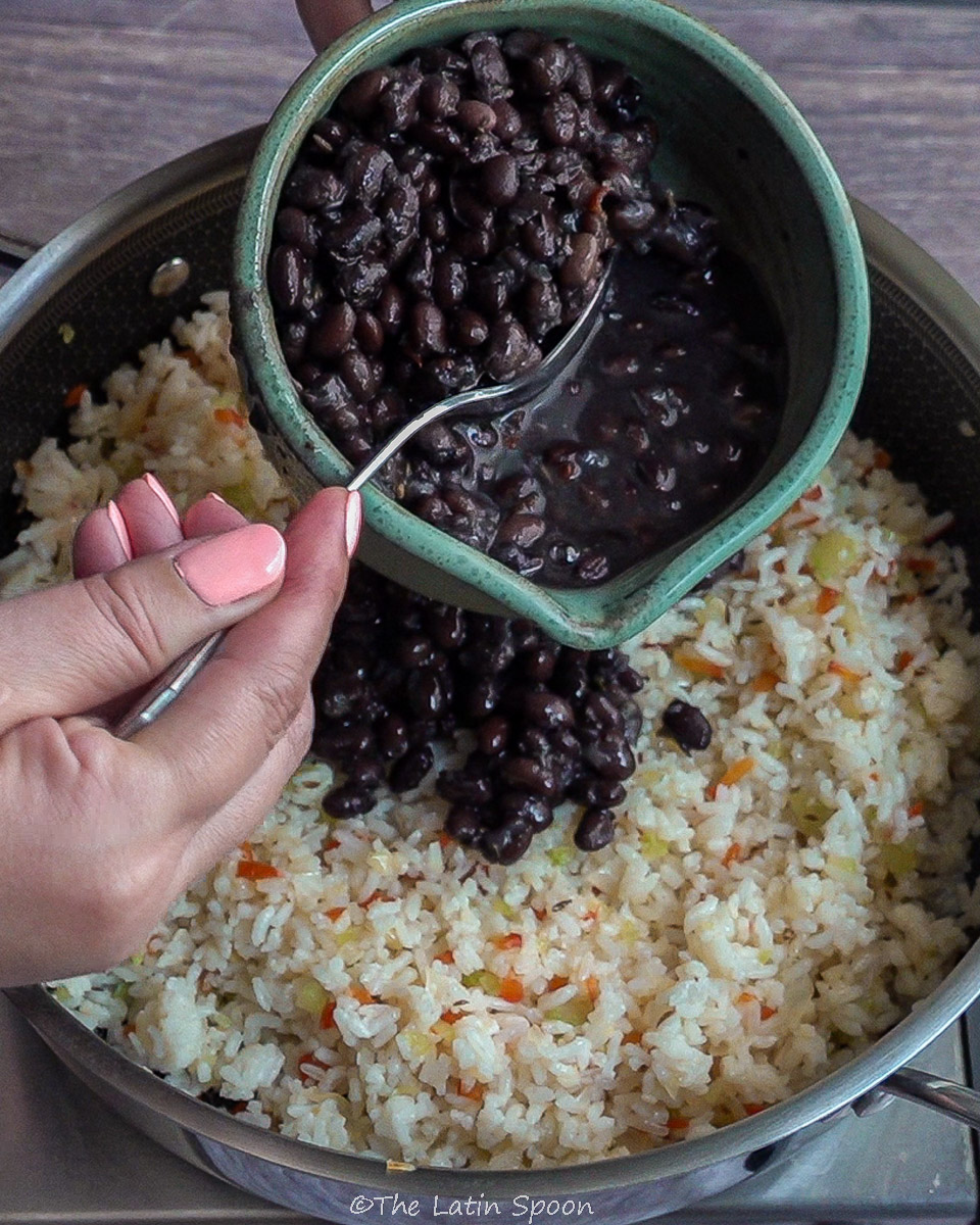 Pan with rice stirred together with sofrito. Above, two hands hold a bowl of beans—the right hand grabbing beans—showing the next step of adding them to the rice.