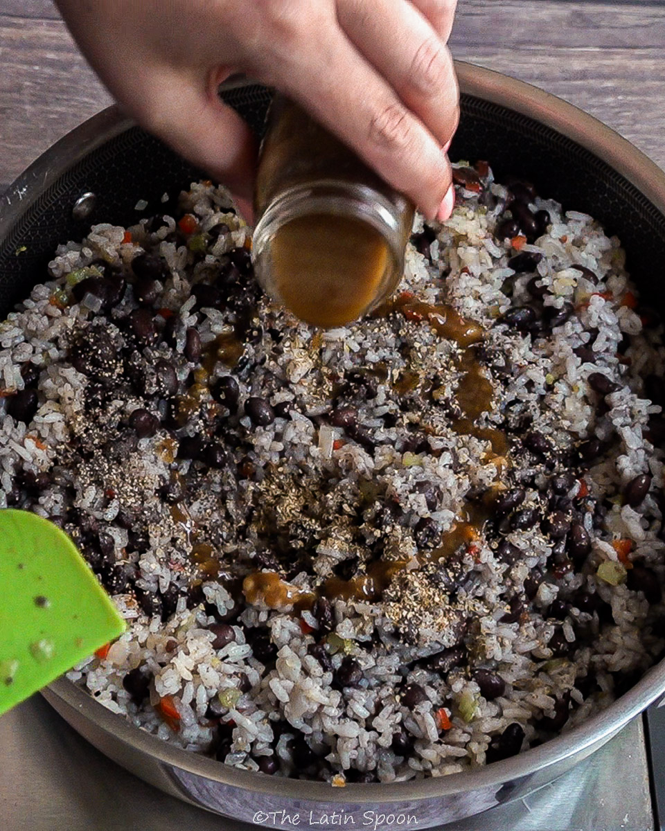 Pan filled with rice and beans mixed together, while a left hand adds Salsa Lizano to the mixture. Oregano and black pepper are already visible on the rice and beans.