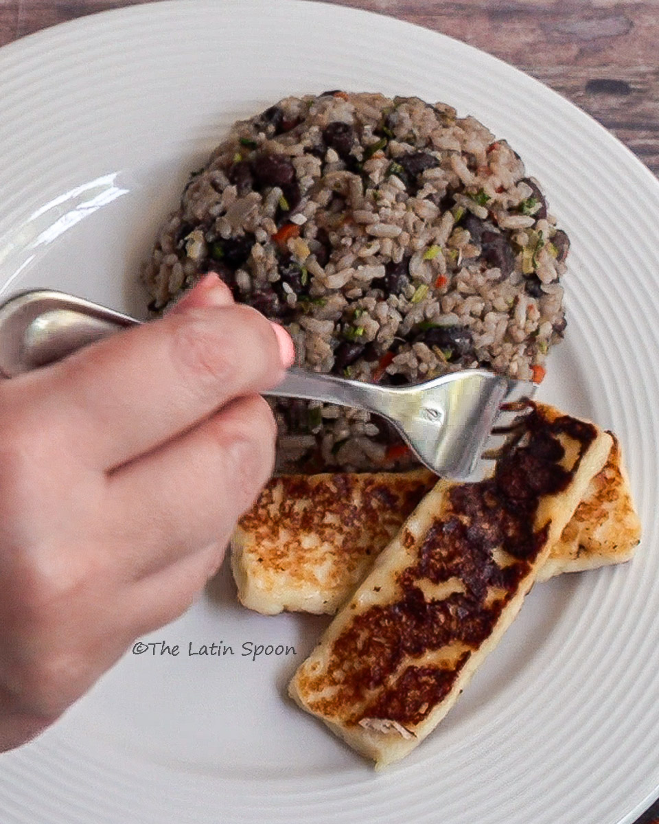 White round plate with Gallo Pinto neatly shaped into a mound. Right hand adds a piece of fried cheese to the plate. The cheese is rectangular and golden brown.