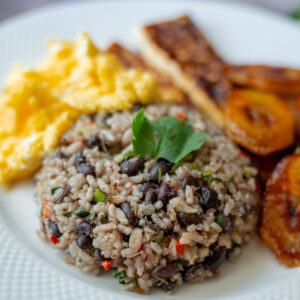 White plate beautifully arranged with Gallo Pinto, scrambled eggs, fried plantains, and fried cheese. The Gallo Pinto is topped with a cilantro leaf and shown from the front angle.