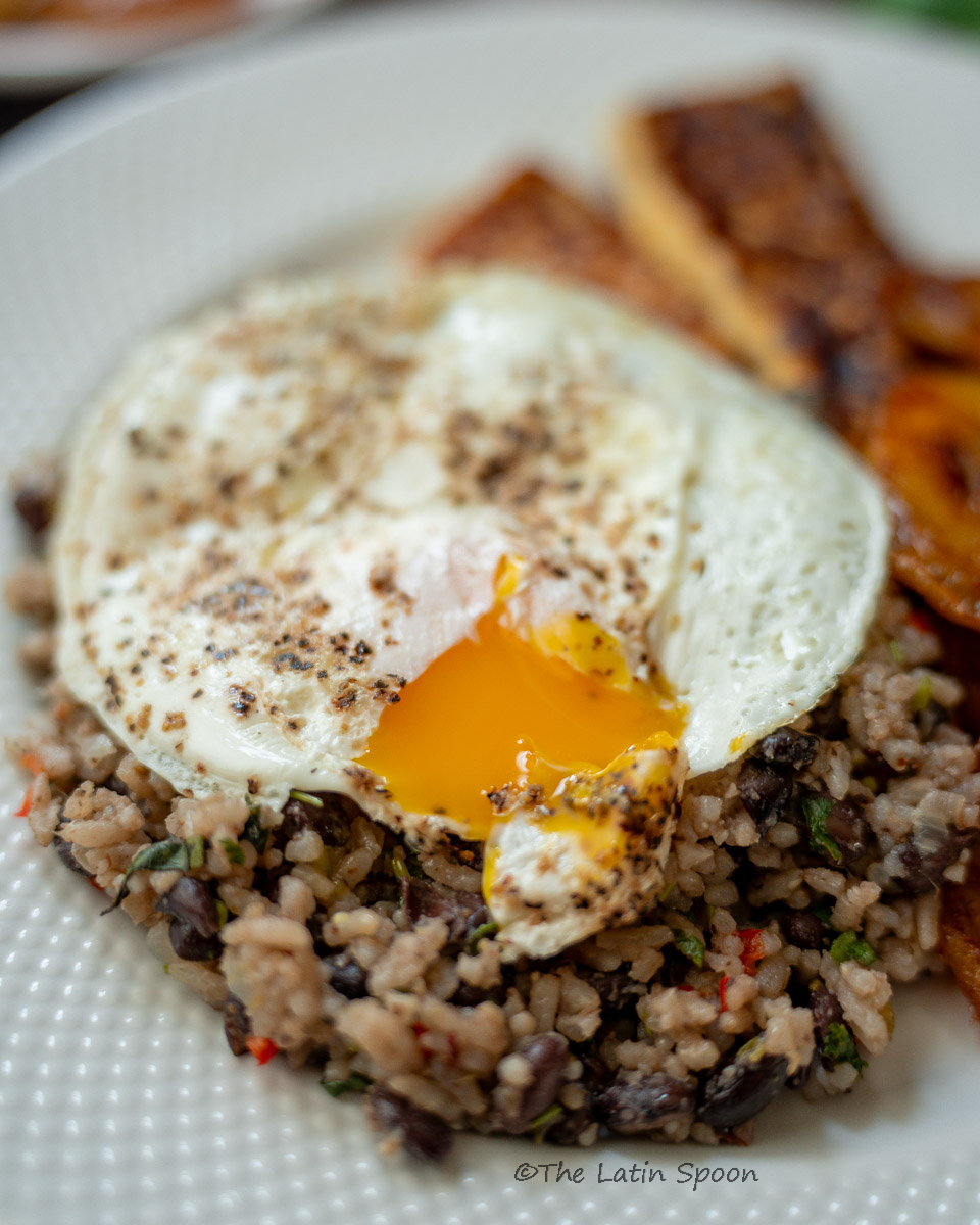 White plate with Gallo Pinto topped with a sunny-side-up egg, the runny yolk spreading throughout the rice and beans.
