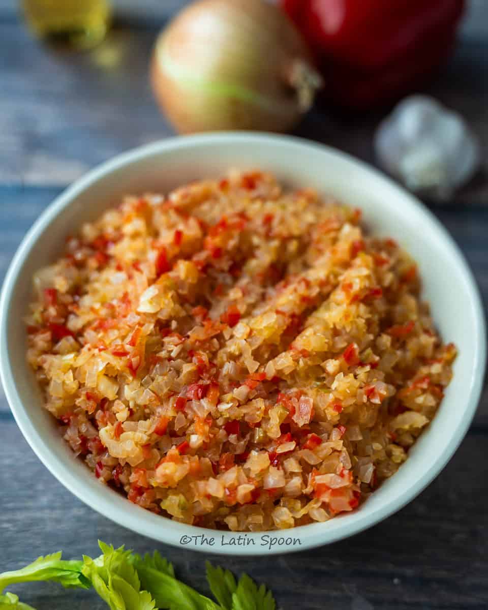 White round bowl filled with cooked sofrito; behind it, a blurred onion, red bell peppers, and garlic can be seen, while celery sticks are placed in front.