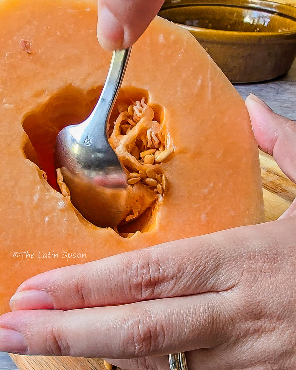 Half cantaloupe held in the left hand while the right hand uses a spoon to scoop out the seeds.