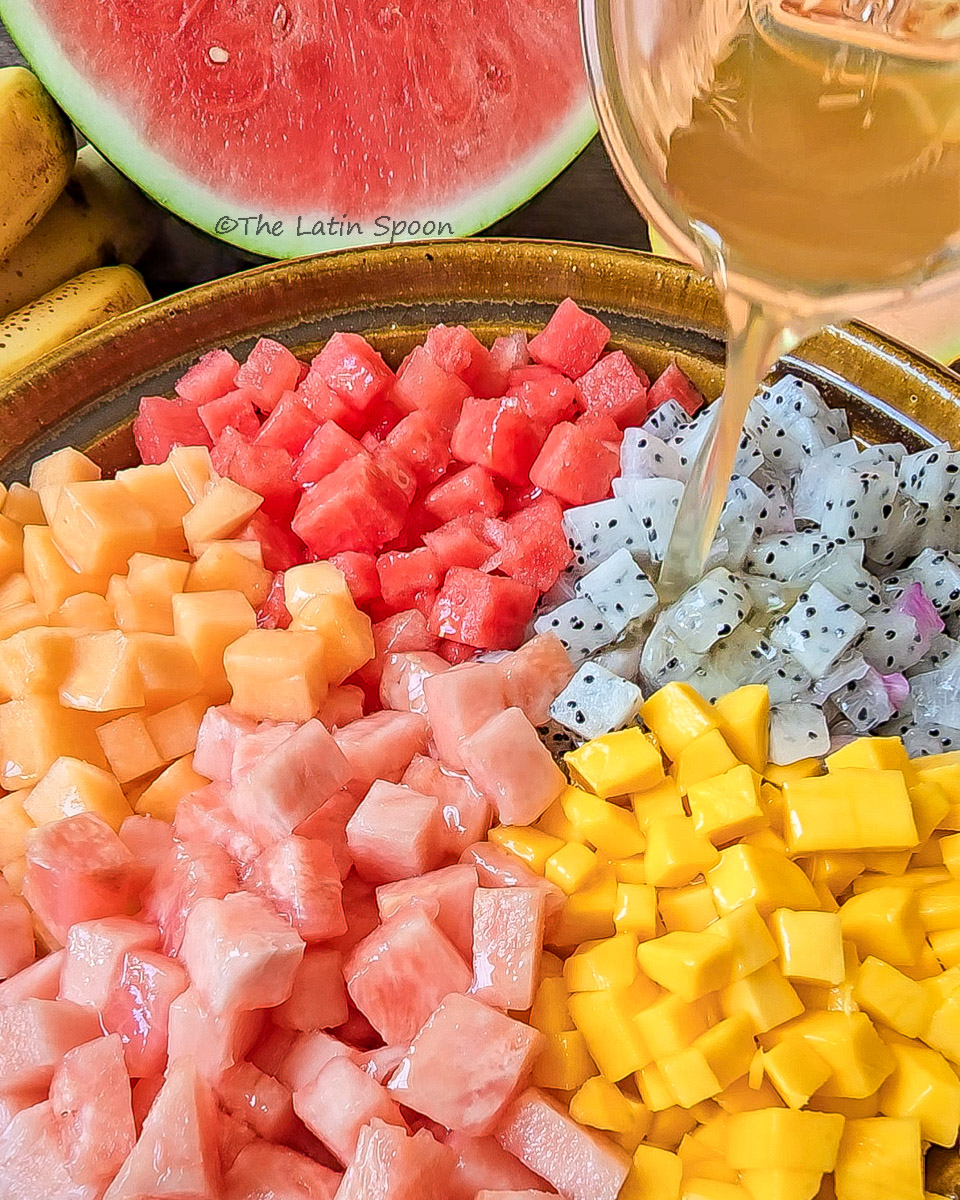 Large bowl of assorted tropical fruits with a small cup pouring honey and lemon juice mixture.
