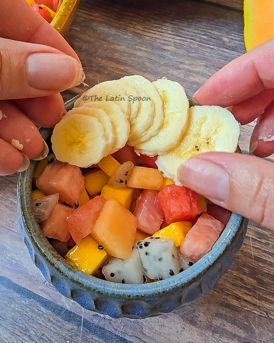 Small bowl of tropical fruit salad with two hands adding banana slices on top.