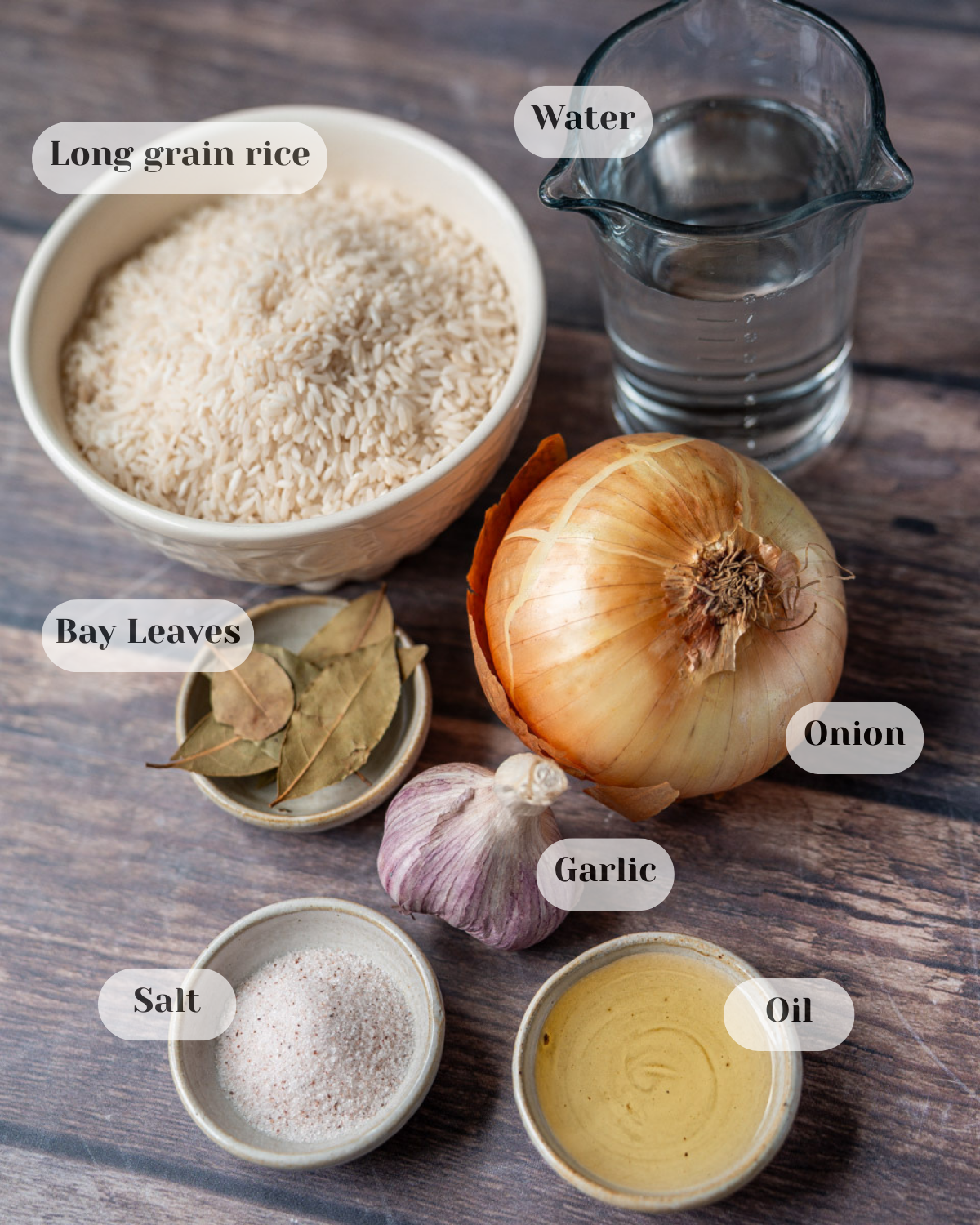 Head shot of all ingredients placed on a wooden table, including rice, garlic, oil, water, onion, salt, and bay leaves