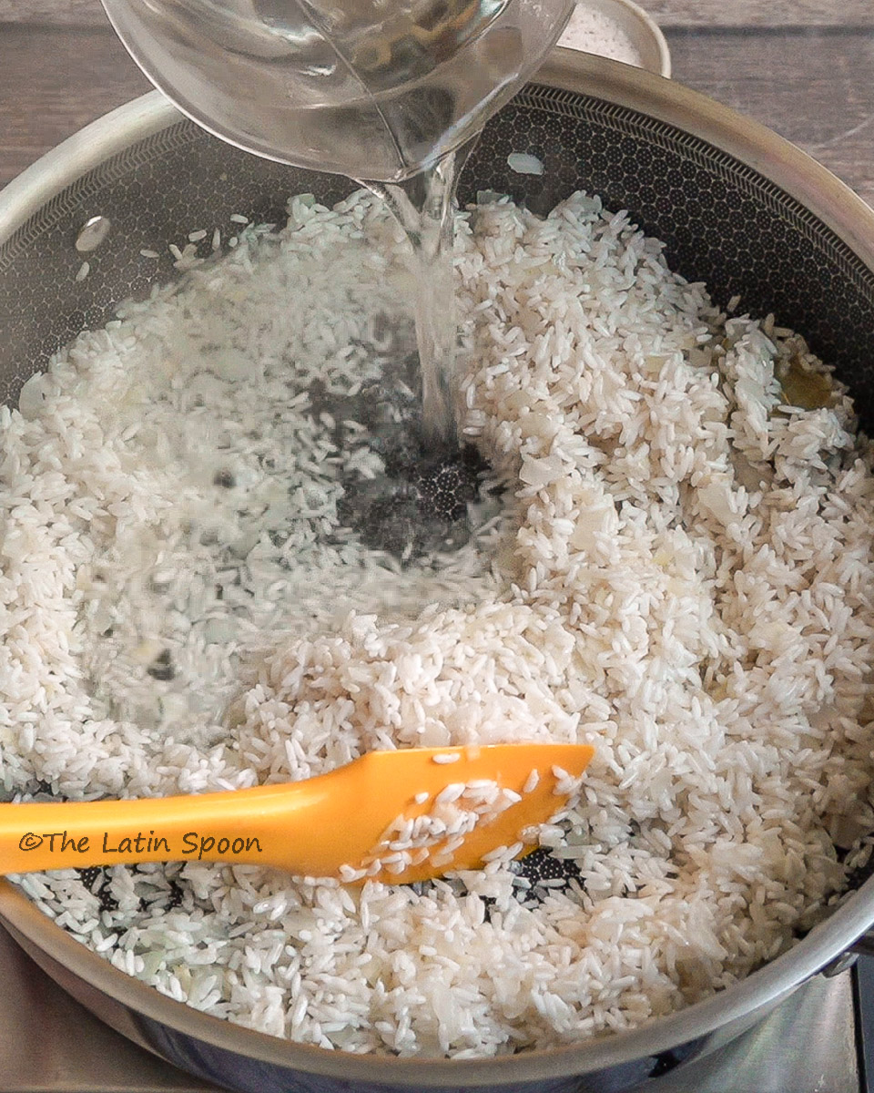 Hot water being poured into rice mixed with garlic, onions, and oil in the pan