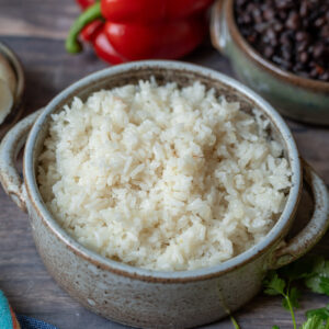 A round ceramic pot filled with fully cooked rice, surrounded by red bell peppers, a small pot of black beans, and fresh cilantro leaves