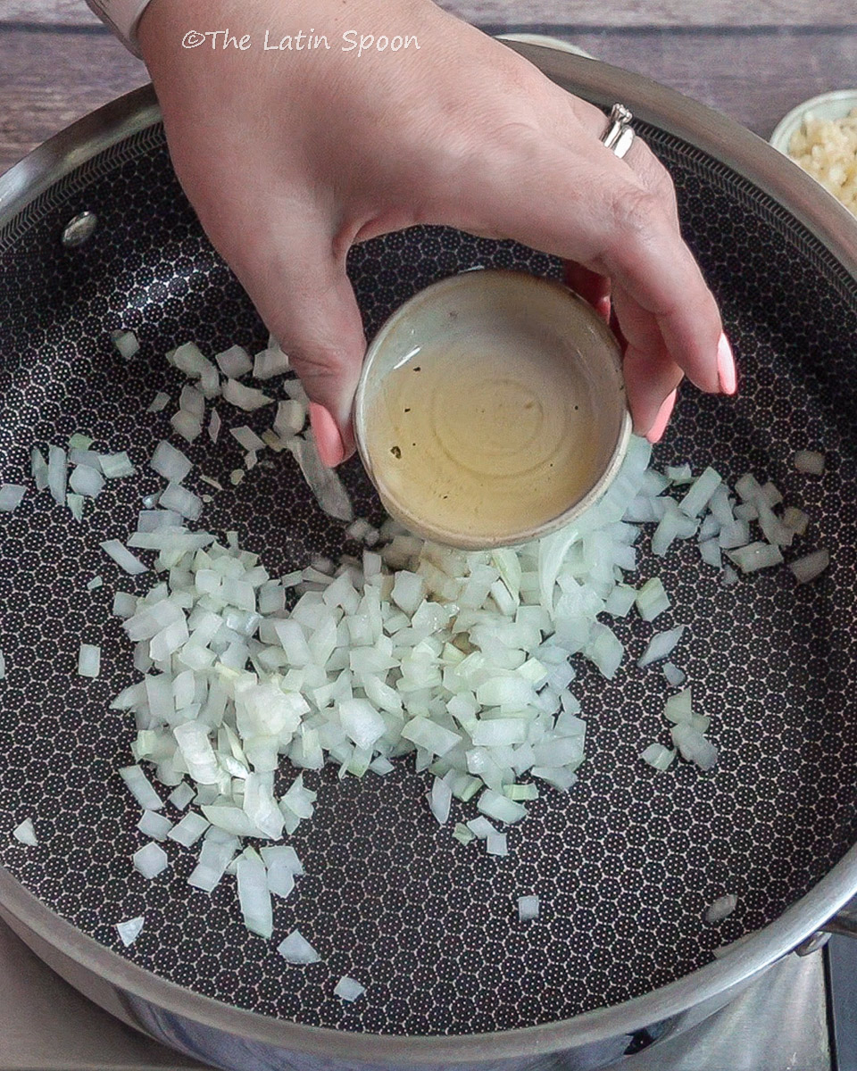 Chopped onions cooking in a large pan while a left hand adds a drizzle of oil