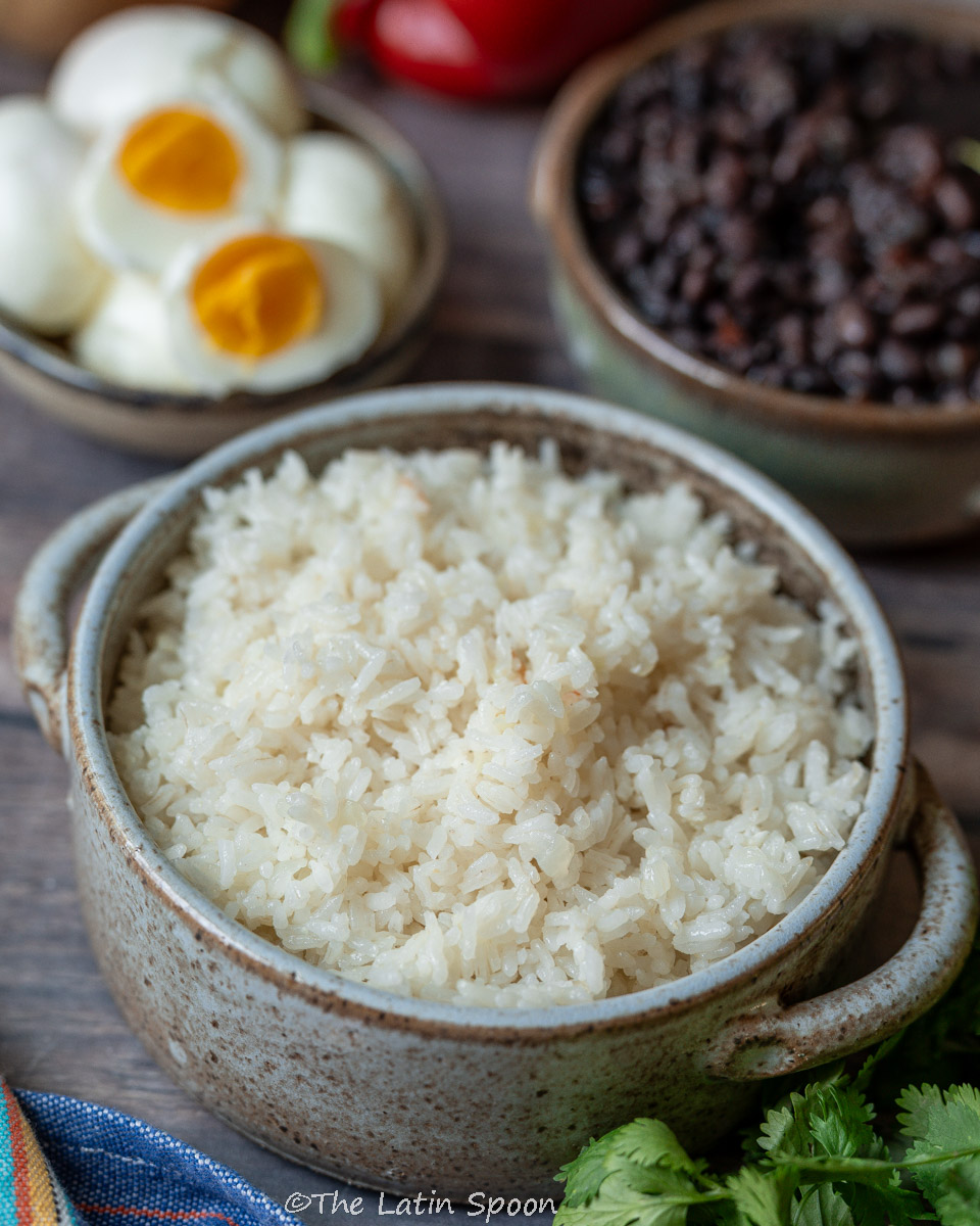 A round ceramic pot filled with fully cooked rice, surrounded by red bell peppers, a small pot of black beans, and fresh cilantro leaves