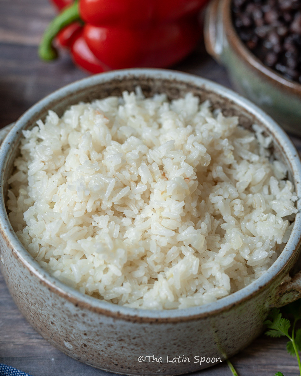 A round ceramic pot filled with fully cooked rice, surrounded by red bell peppers, a small pot of black beans, and fresh cilantro leaves