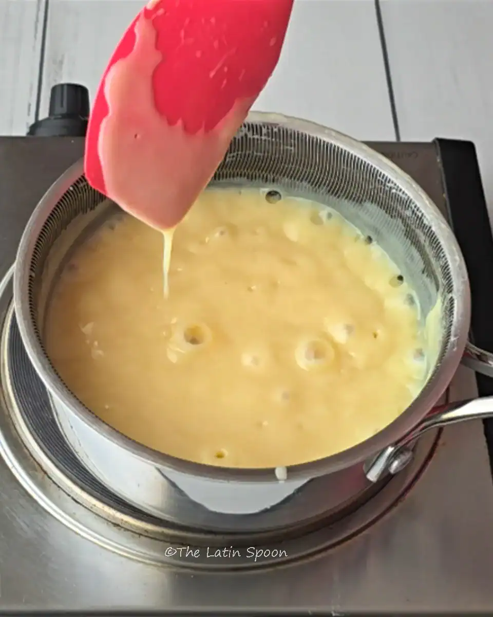 A small pot cooking the condensed milk frosting for the cake, with small bubbles showing it is thickening over medium heat, and a pink spatula dripping a bit to show the consistency.
