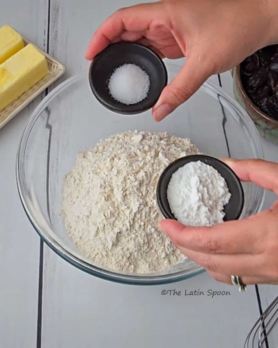A clear glass bowl with flour at the bottom, and two hands above it, the right hand holding a small cup of salt and the left hand holding a small cup of baking powder.