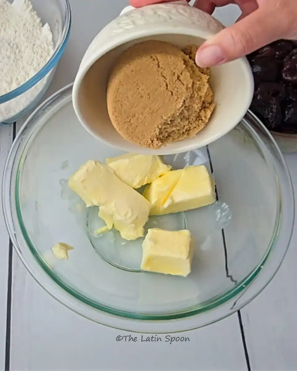 A clear glass bowl containing medium-sized cubes of butter with brown sugar being poured in.