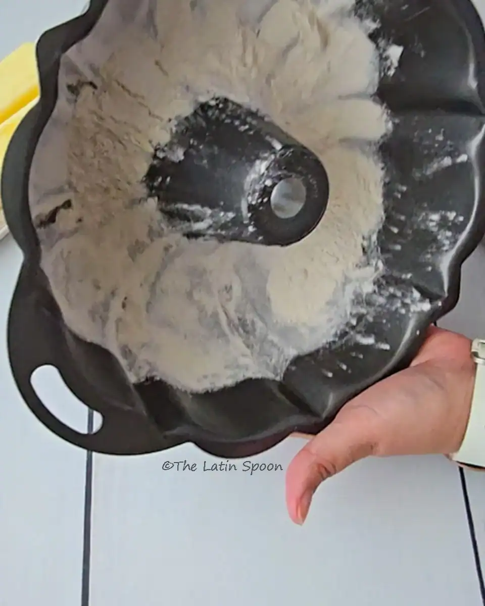 A bundt cake pan seen from the side, with the left hand holding and rotating the pan to spread the flour evenly on the sides.