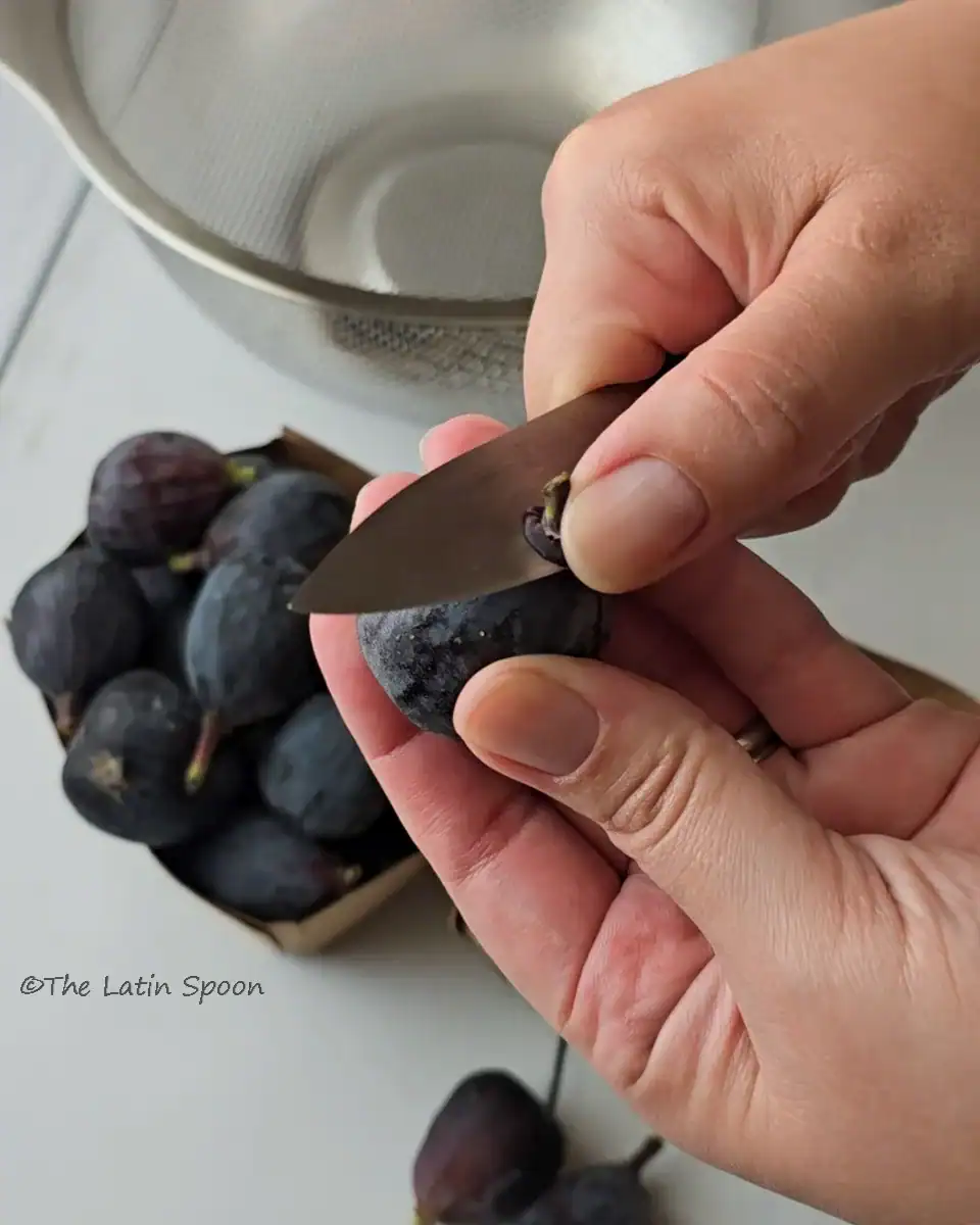 Two hands holding figs, with the right hand cutting off the stems using a knife.