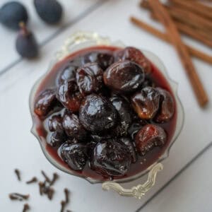 Overhead view of fig preserves in their syrup, with cinnamon sticks, fresh figs, and whole cloves visible in the background.