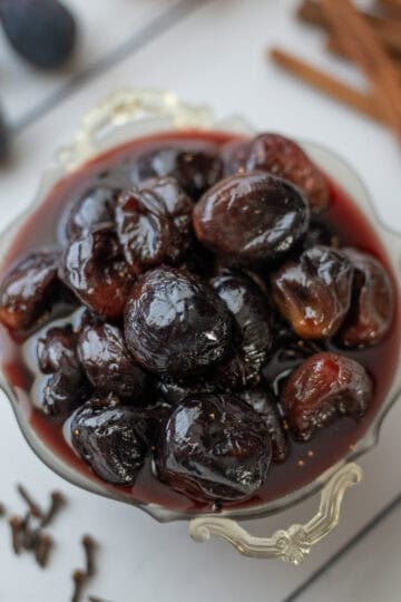 Overhead view of fig preserves in their syrup, with cinnamon sticks, fresh figs, and whole cloves visible in the background.