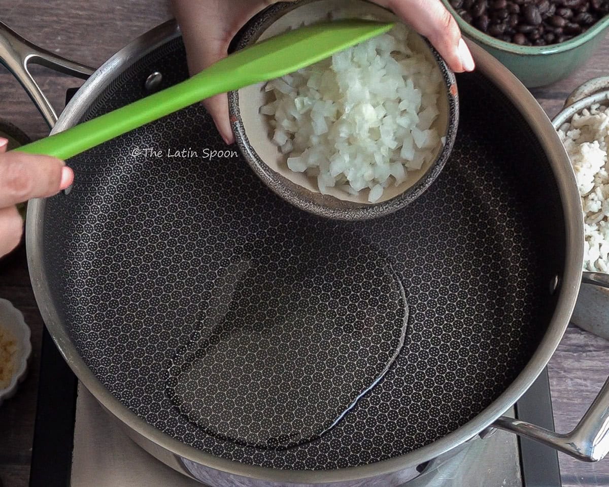 Large saucepan or skillet with a little oil. Above it, a left hand holds a small bowl of onionacs, being pushed out of the bowl with a green spatula.