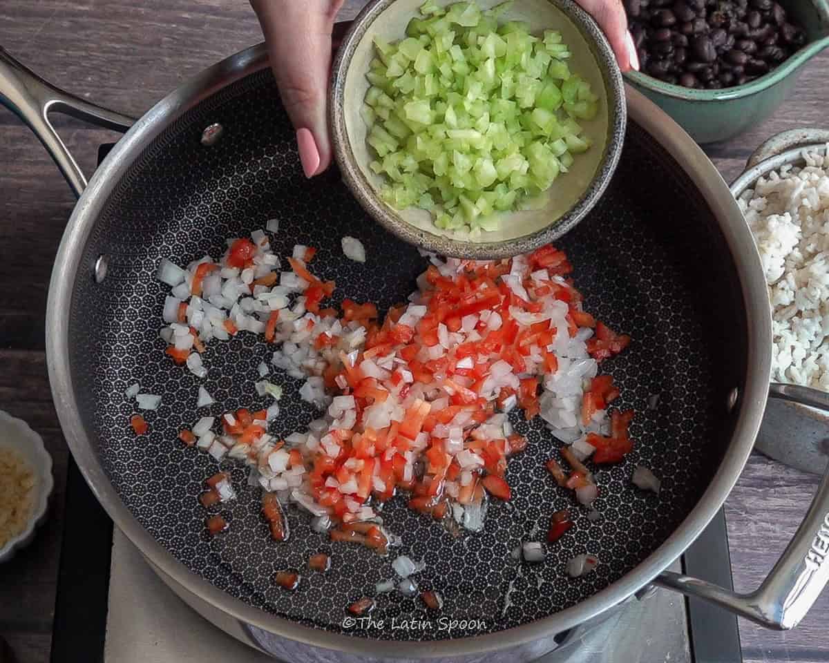Large pan with chopped onions and bell peppers cooking in oil, while a left hand holds a small bowl of chopped celery, showing the next step of adding celery to the pan.