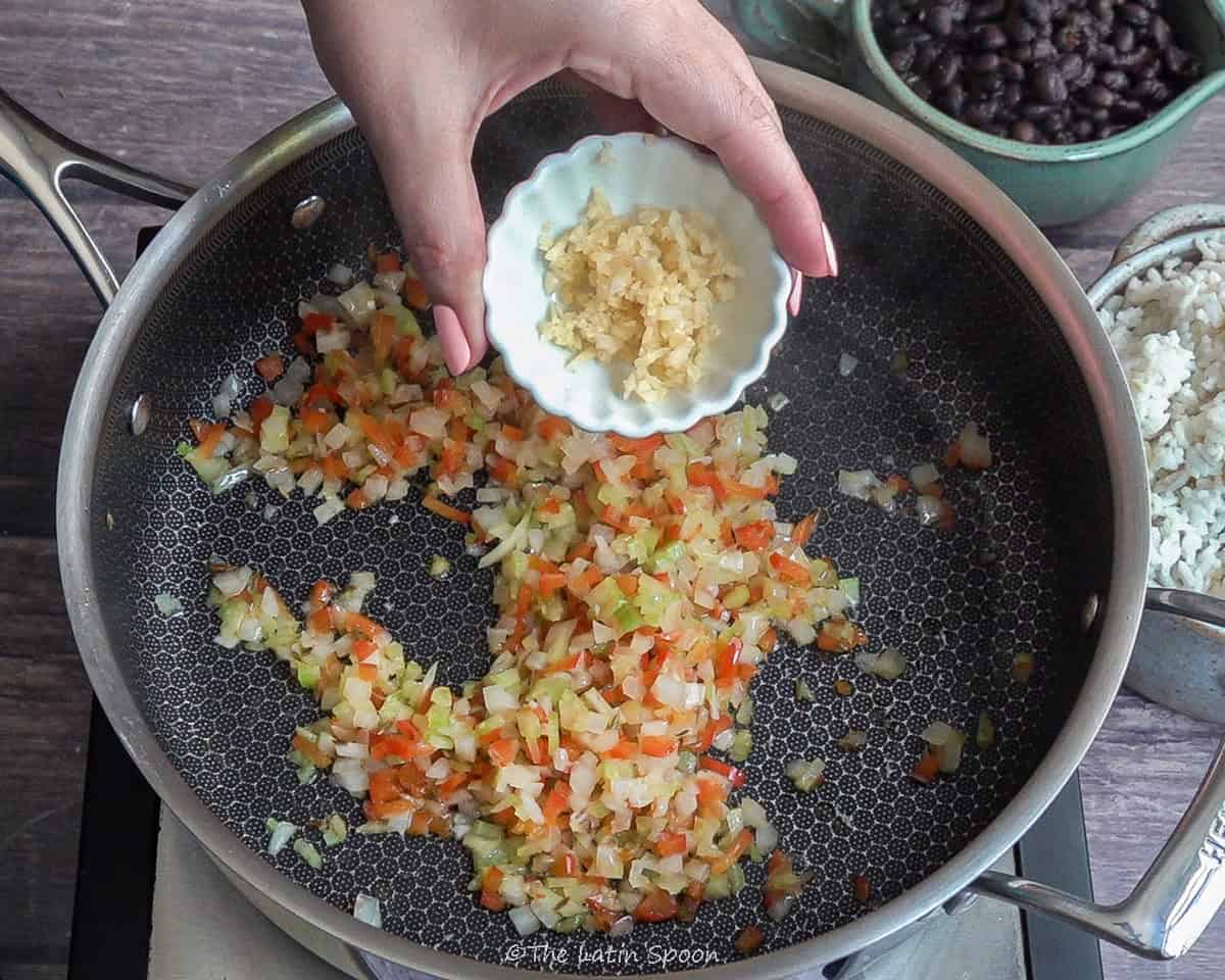 Pan with sofrito (onions, celery, bell peppers, and oil) cooking, while the left hand is about to add minced garlic from a small bowl.