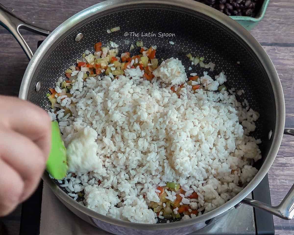 Large pan showing sofrito at the bottom with fully cooked white rice added on top. In the corner, a green spatula begins to mix the ingredients.