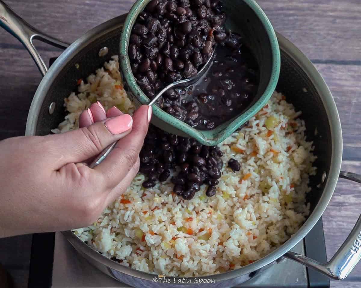 Pan with rice stirred together with sofrito. Above, two hands hold a bowl of beans—the right hand grabbing beans—showing the next step of adding them to the rice.