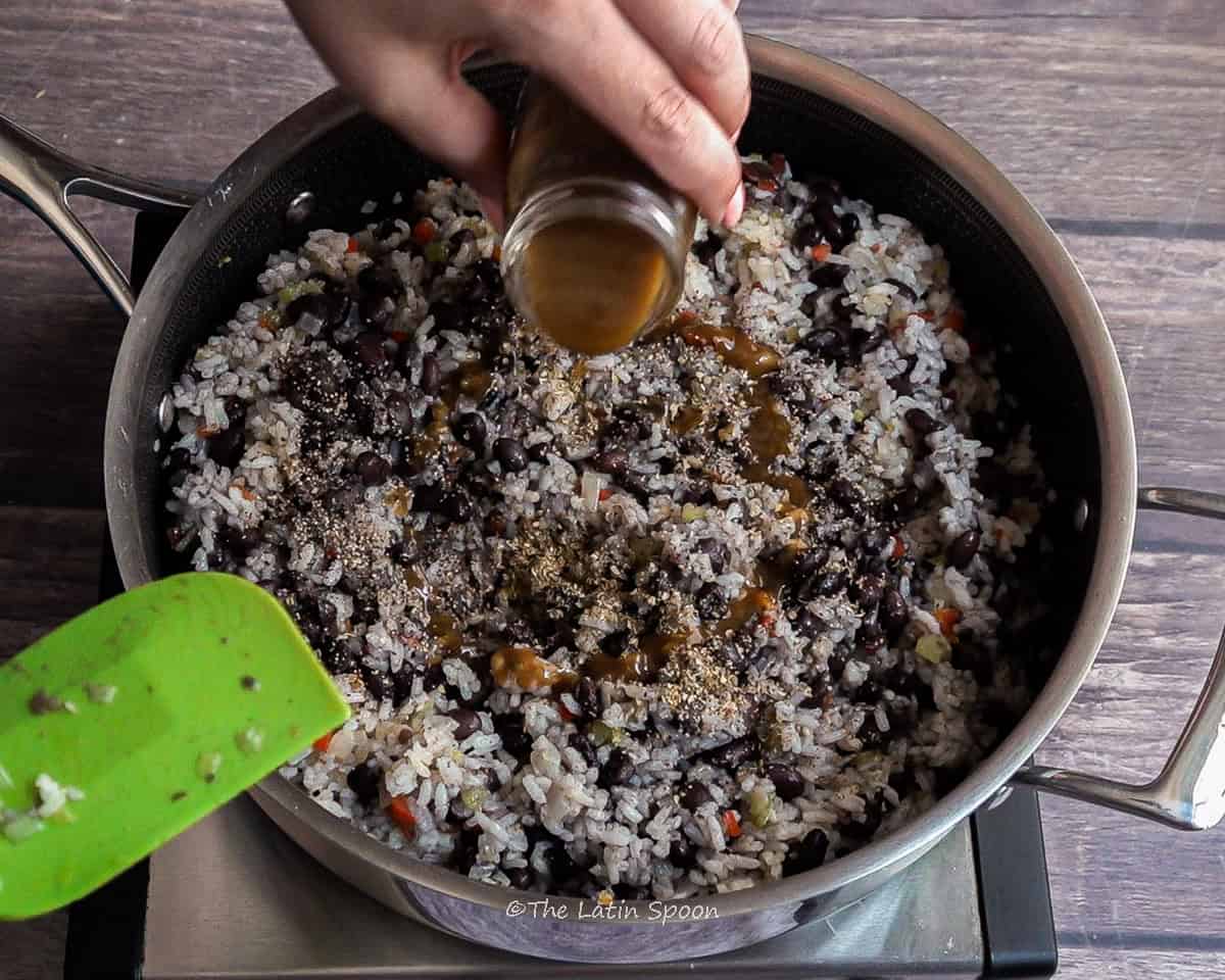 Pan filled with rice and beans mixed together, while a left hand adds Salsa Lizano to the mixture. Oregano and black pepper are already visible on the rice and beans.