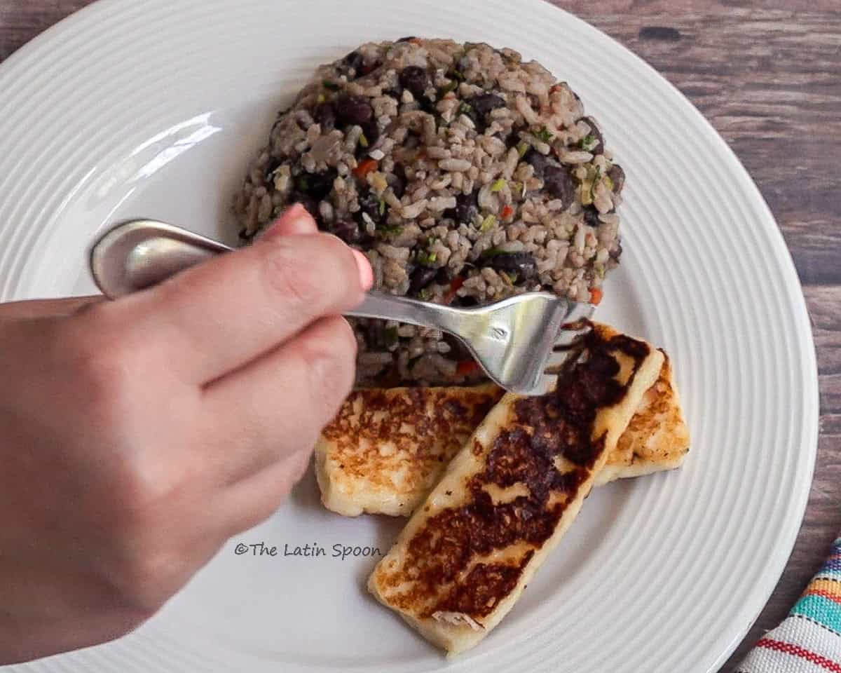White round plate with Gallo Pinto neatly shaped into a mound. Right hand adds a piece of fried cheese to the plate. The cheese is rectangular and golden brown.
