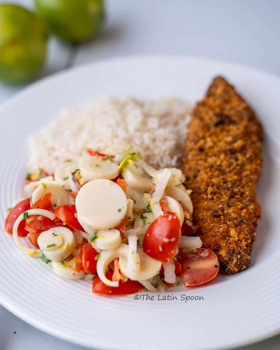 A white plate with a finished dish ready to enjoy, featuring white rice, heart of palm salad, and a breaded tilapia fillet.