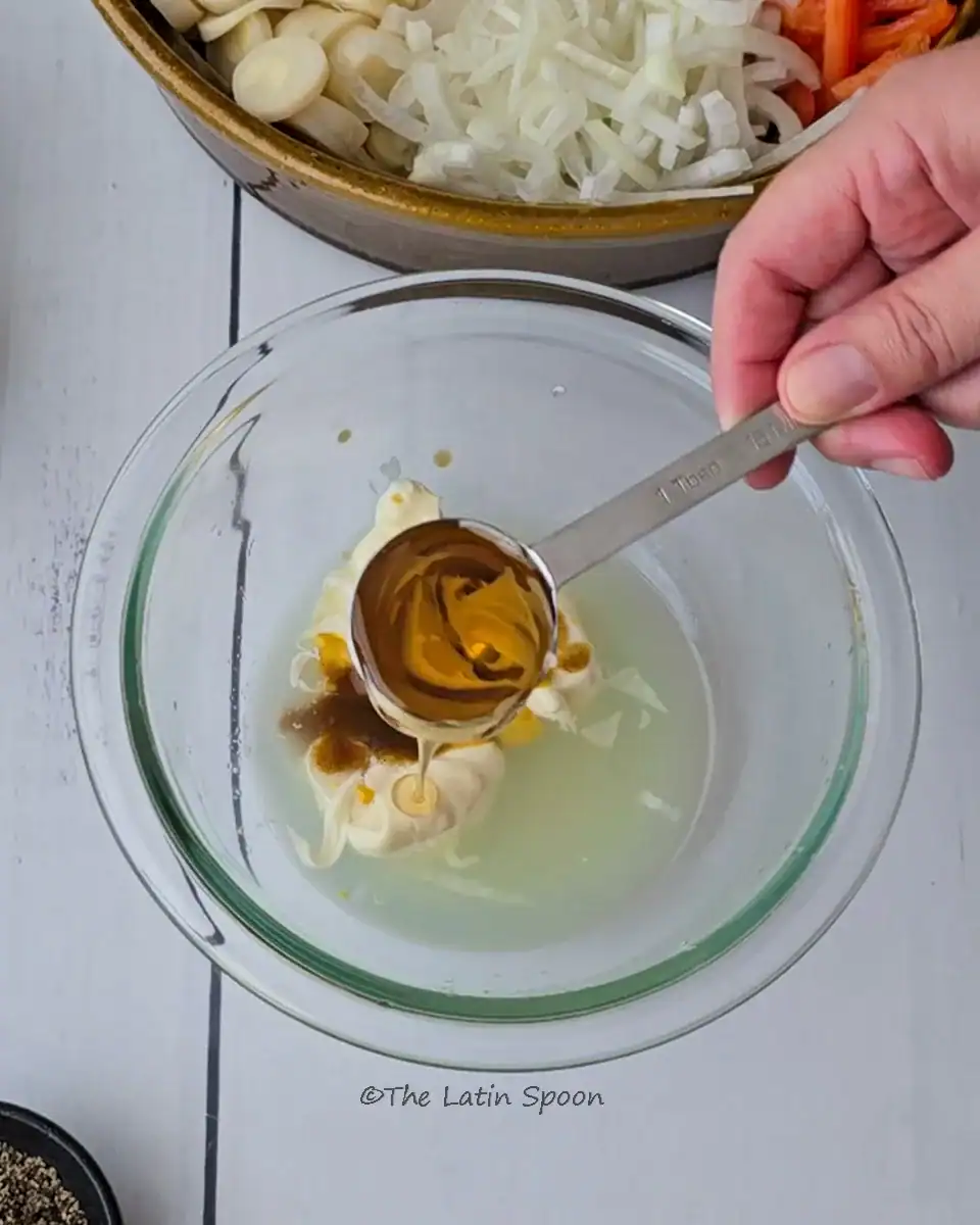 A medium clear glass bowl with lemon juice and mayonnaise at the bottom, and a spoon adding agave syrup.