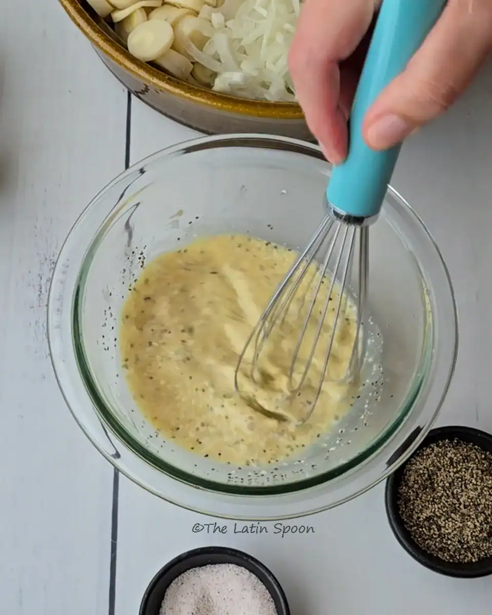 A small clear glass bowl with liquid ingredients inside and a whisk mixing what appears to be the salad dressing.