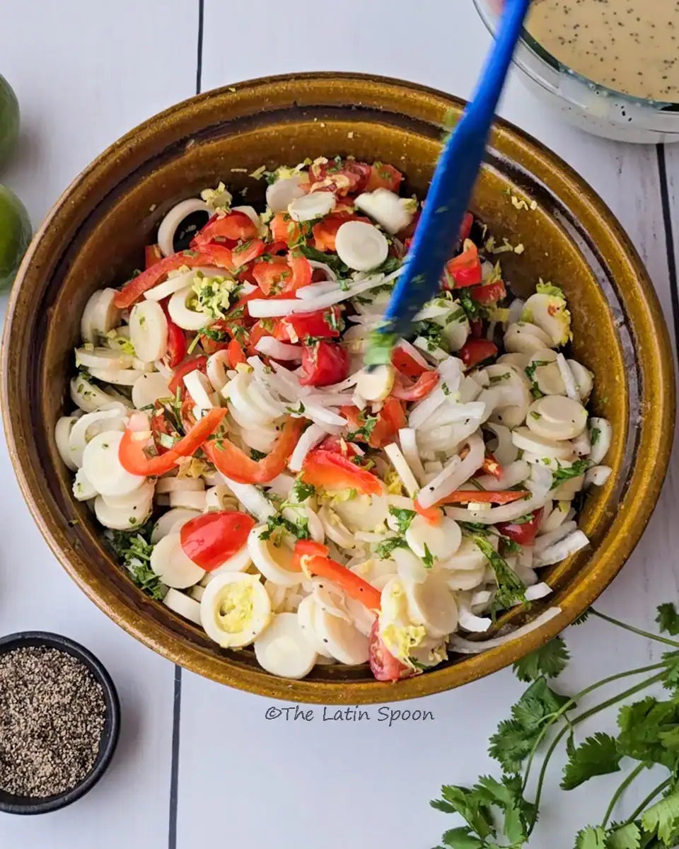 A brown bowl with heart of palm salad being mixed with a blue spatula, with a small cup of black pepper and fresh cilantro visible outside the bowl.