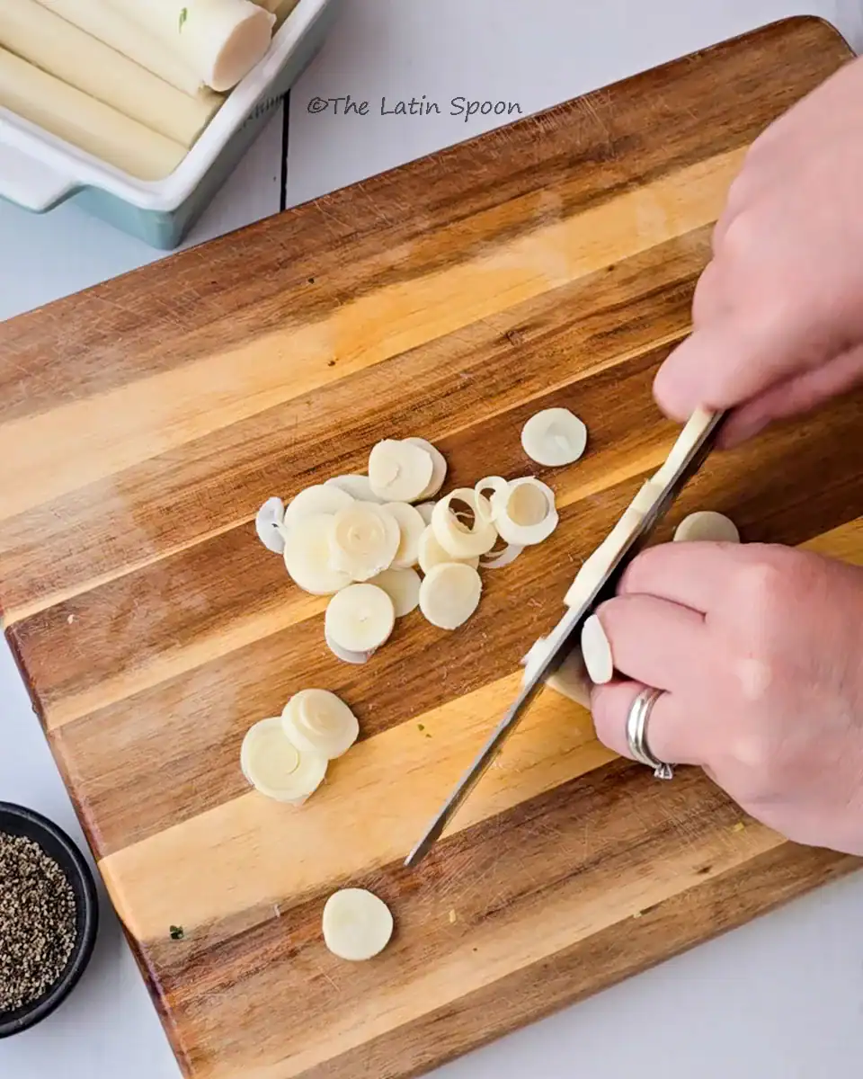 A wooden cutting board with two hands cutting heart of palm, with sliced pieces already on the board.
