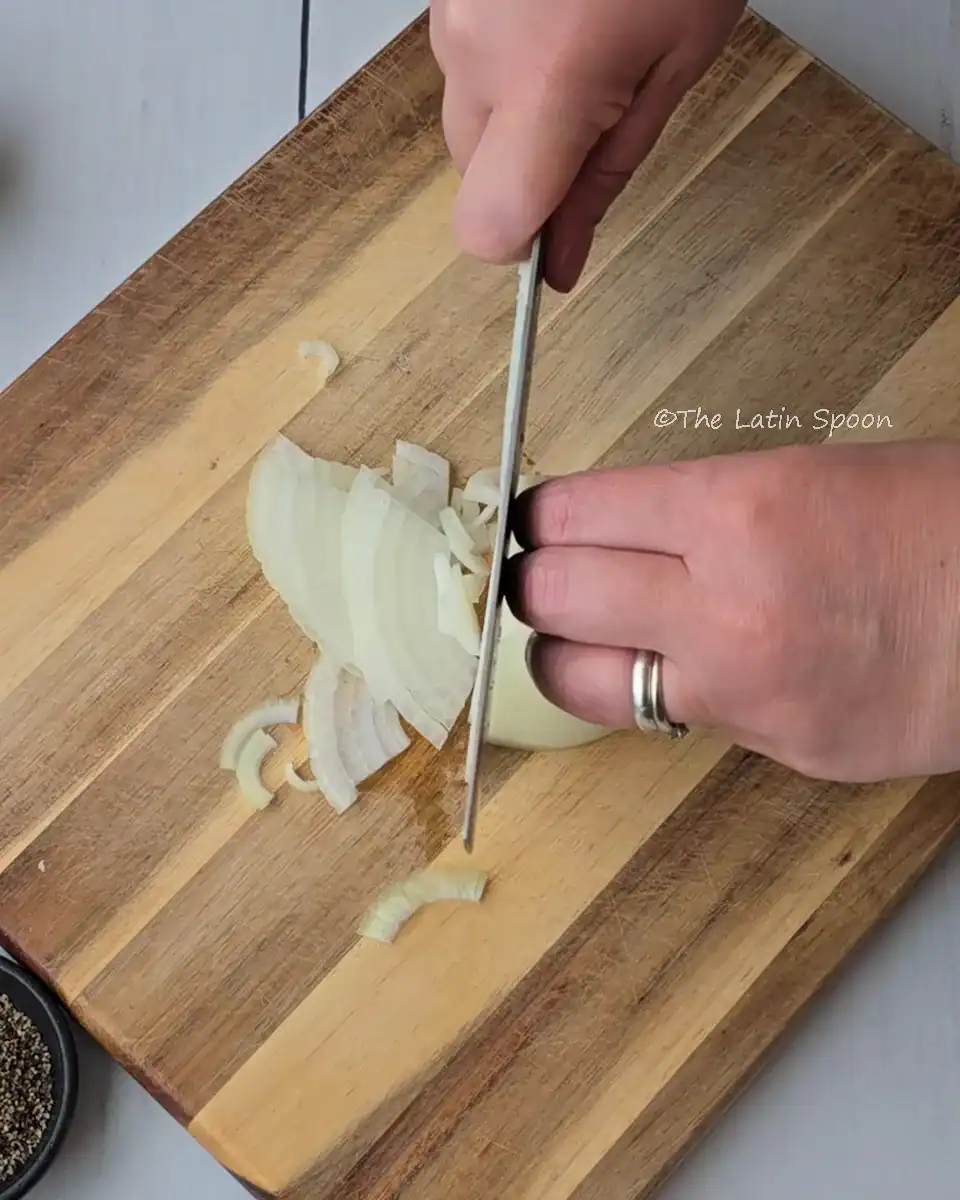 A wooden cutting board with a white onion being cut, the left hand holding the onion and the right hand cutting it with a knife.