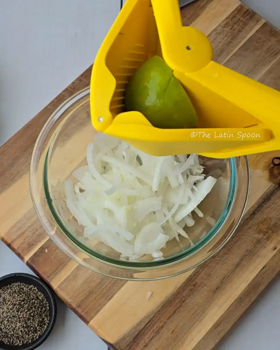 A wooden cutting board with a small glass bowl of onions, and above it a yellow lemon squeezer juicing a green lemon.