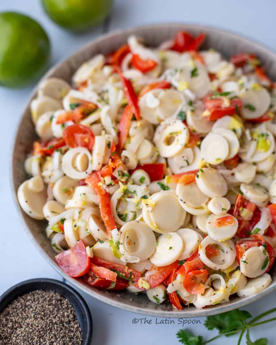 A brown bowl holding the heart of palm salad, showing pieces of heart of palm, tomato, chili, and other ingredients, with green lemons and a small cup of black pepper visible outside the bowl.
