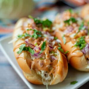 Side view of a Costa Rican hot dog in a bun topped with cabbage, pink sauce, and cilantro. Two hot dogs are on the same square plate with cabbage in the background.
