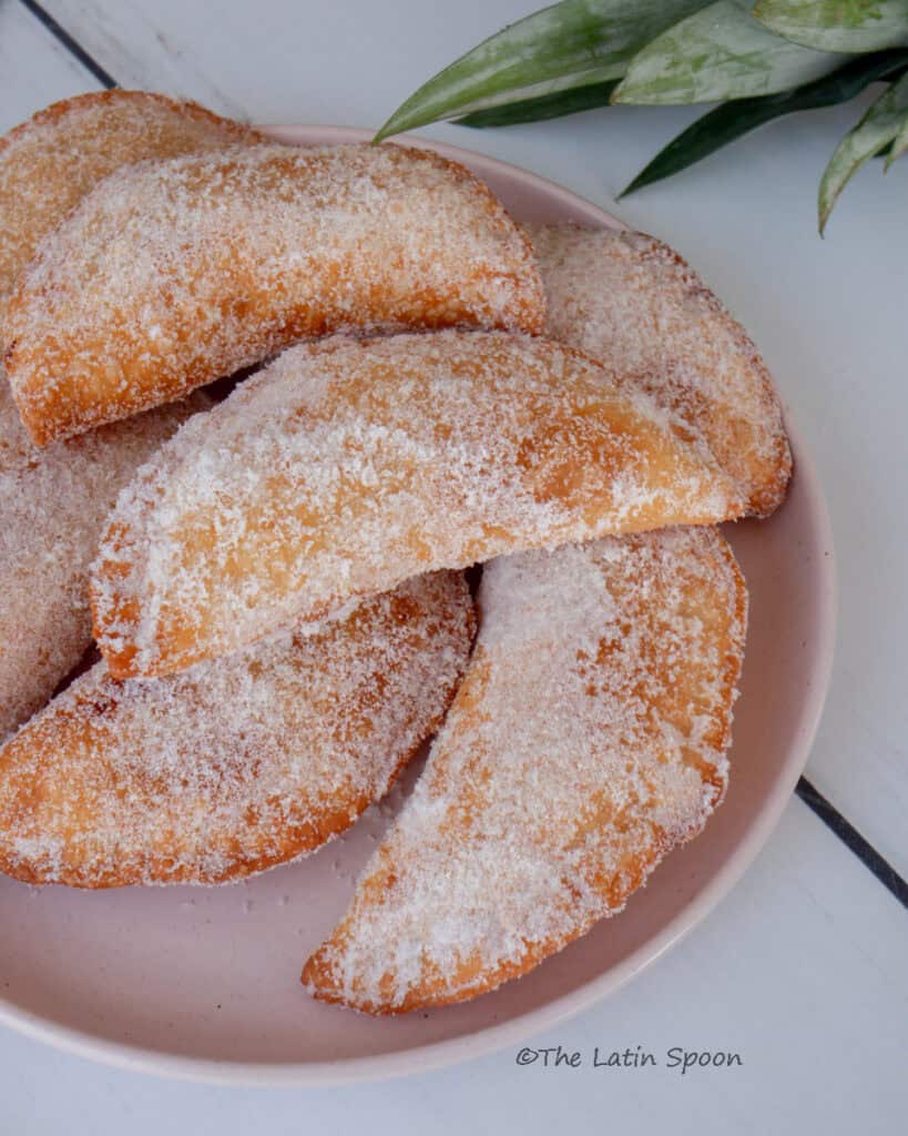 Golden fried empanadas stacked on a pink plate, dusted with sugar.
