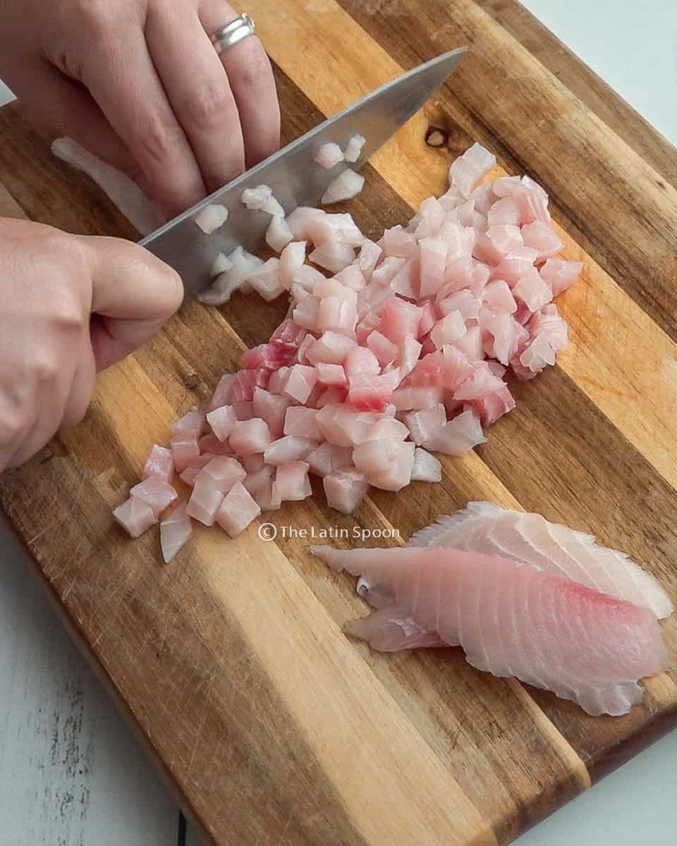On a cutting board, tilapia is being cut into small cubes with a knife