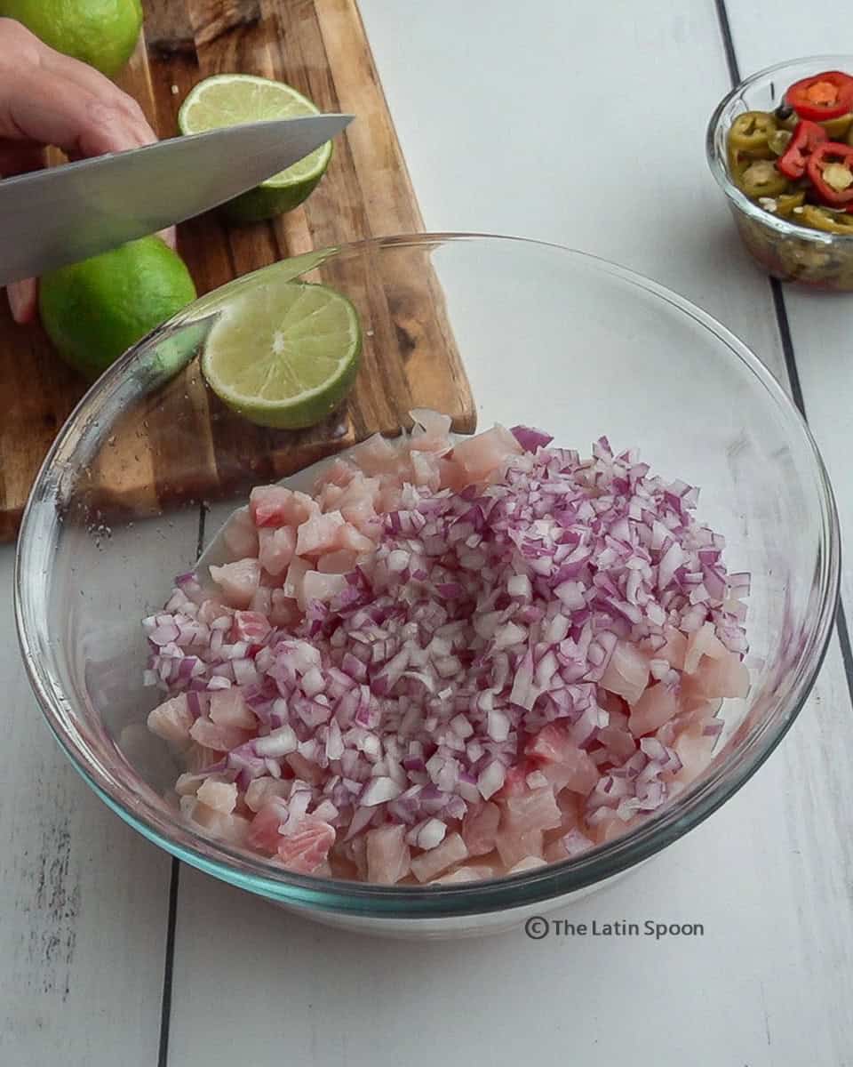 In the foreground, a clear bowl contains pieces of fish and chopped onion. In the background, hands are cutting lemons in half on a cutting board.