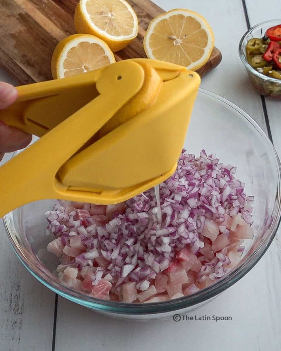 A clear bowl filled with small diced fish and onion, with a lemon squeezer positioned above it as juice drips down