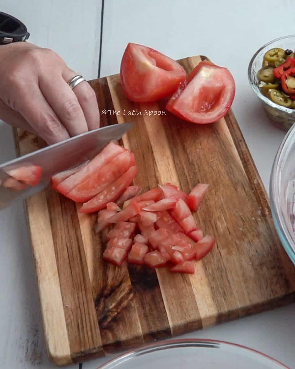 On a cutting board, hands are cutting a tomato into pieces with a knife.