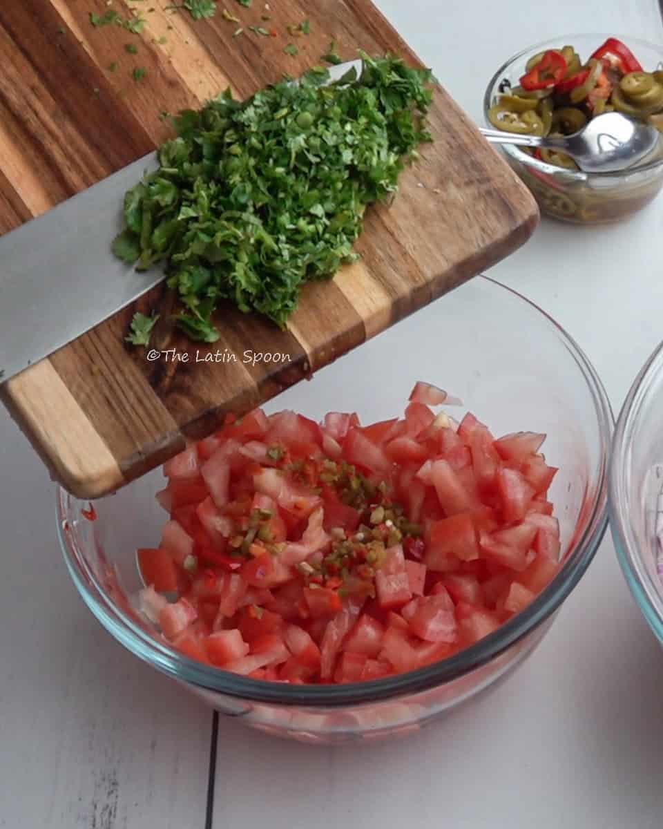 A clear bowl with diced tomatoes and finely chopped jalapeño. Above it, a cutting board is held at an angle while a knife slides chopped cilantro from the board into the bowl.