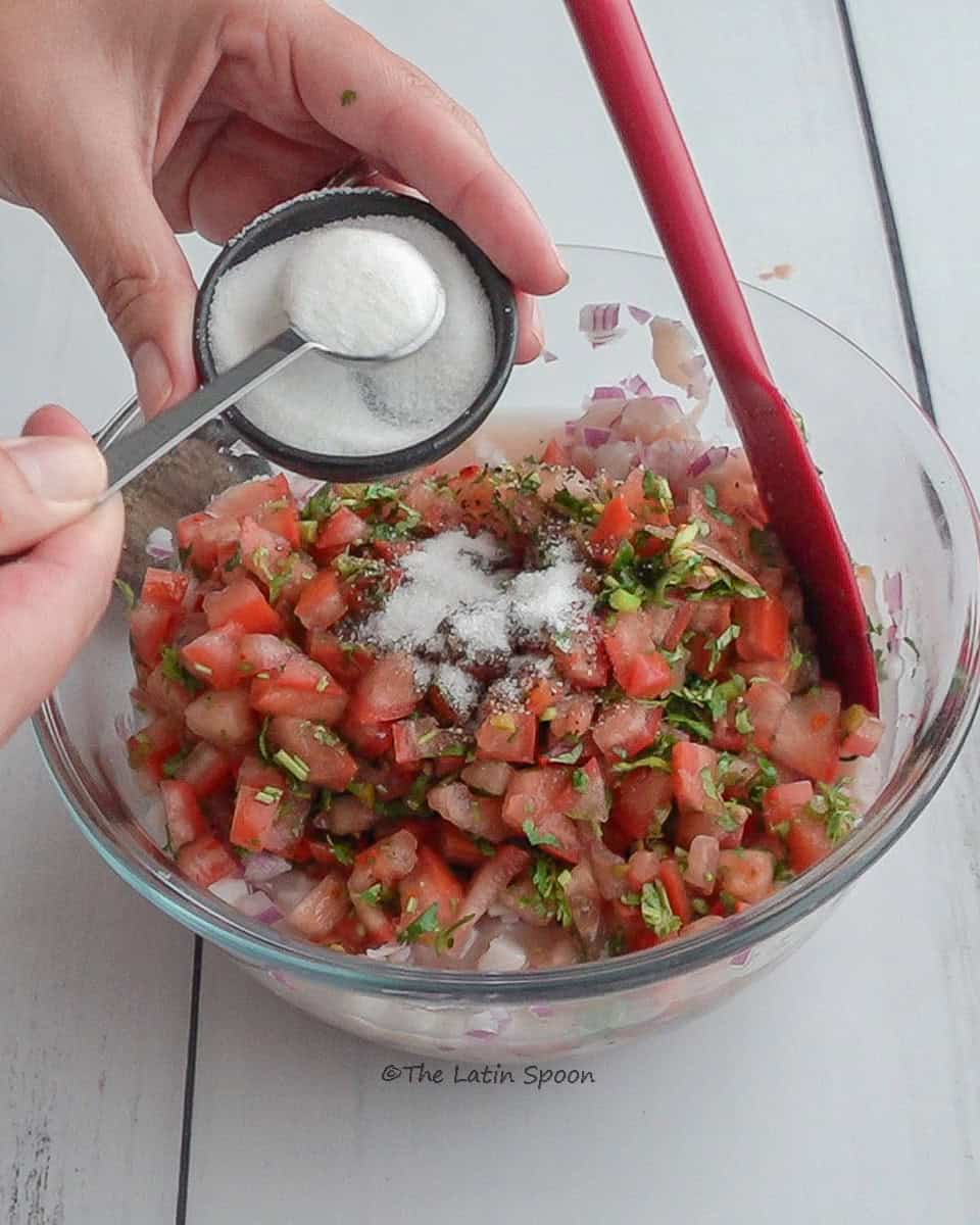 The left hand holds a small bowl with sugar, while the right hand uses a measuring spoon to scoop sugar from the bowl to add it into a clear bowl below that contains the ceviche mixture.
