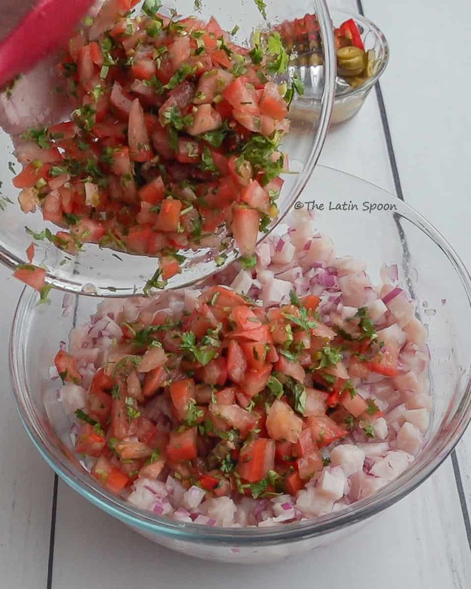 A clear bowl with tomato, cilantro, and jalapeño mixture is being tilted over another bowl on the table that contains diced tilapia, red onion, and lemon juice.